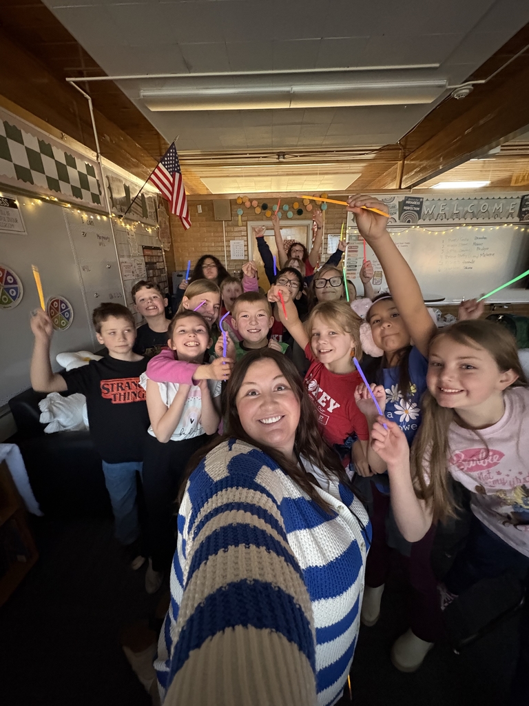 A cheerful group selfie of elementary students and their teacher in a classroom. They are all smiling and holding up colorful, glowing neon sticks.