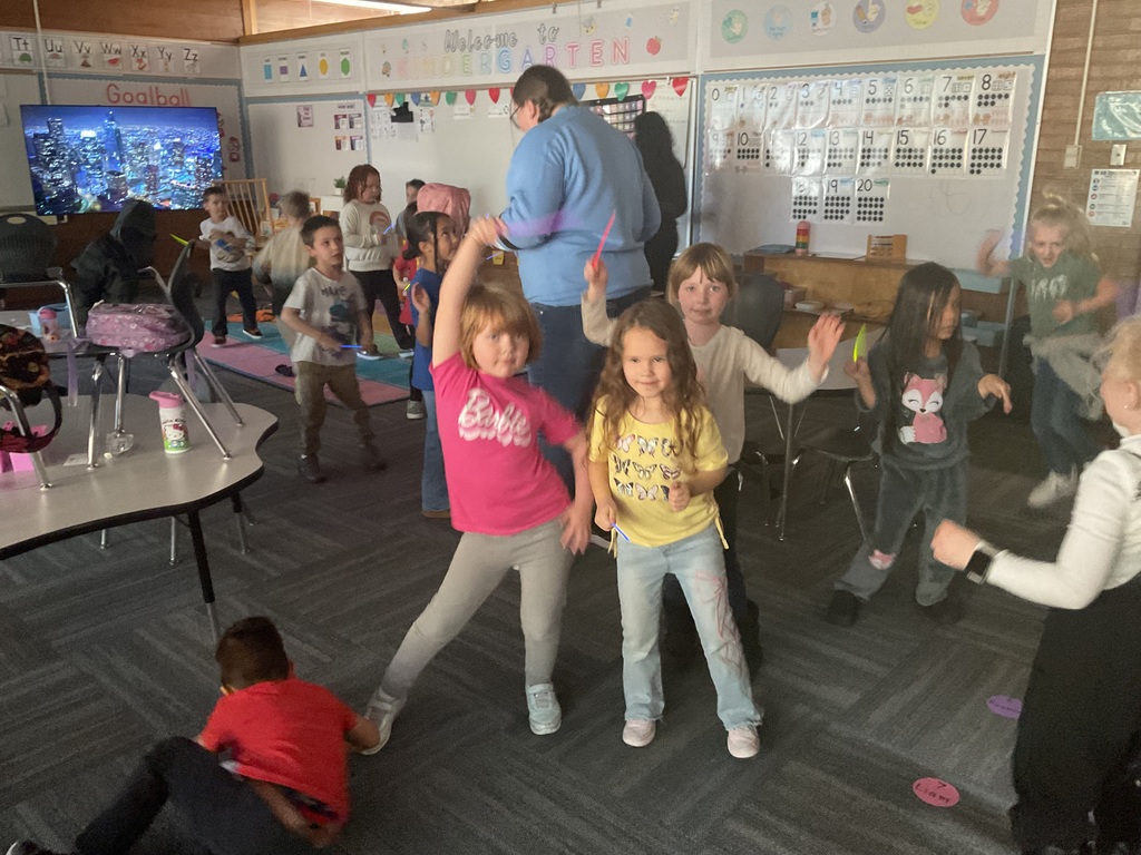 A group of younger students in a "Welcome to Kindergarten" classroom dancing with purple and pink glow sticks. A cityscape is visible on a TV screen in the background.