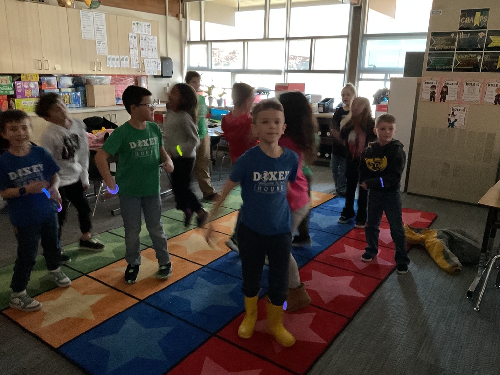 Several students wearing "Doxey" shirts dance and move on a colorful rug featuring large stars. They are wearing glowing neon bracelets and rings while they play.