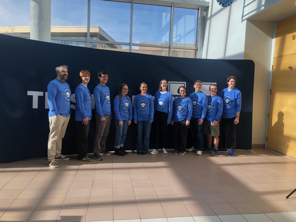 A group of students and one adult coach stand in a line indoors, wearing matching blue Academic Olympiad long-sleeve shirts. They are posing and smiling in front of a dark backdrop inside a bright school building with large windows and natural light.