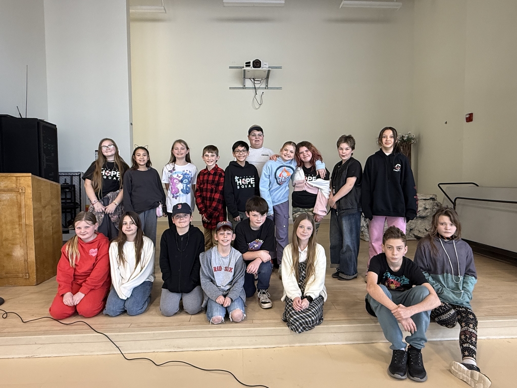 A group of 18 elementary students pose together on a wooden stage after a Geography Bee competition. The students are arranged in two rows, with some kneeling in front and others standing behind them. They are dressed in casual school clothes, including hoodies and t-shirts, and the background shows a simple white wall with a ceiling-mounted projector.