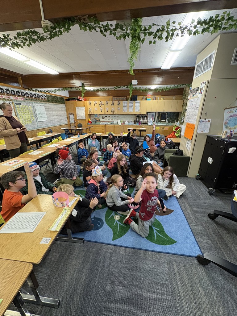A large group of students sits cross-legged on a blue rug featuring a leaf pattern. They are looking toward the front of the classroom, and several students have their hands raised to answer a question.