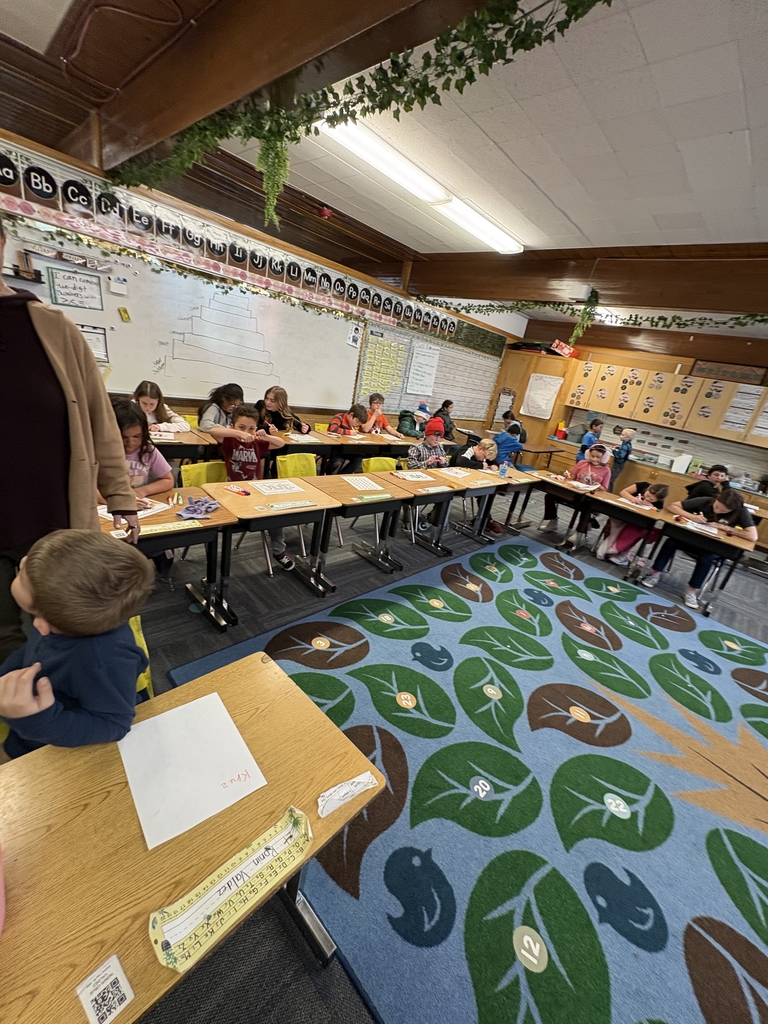 An angled view of the classroom showing students at their desks working on coloring activities and worksheets. The room features wooden cabinetry and a green vine border along the ceiling beams.