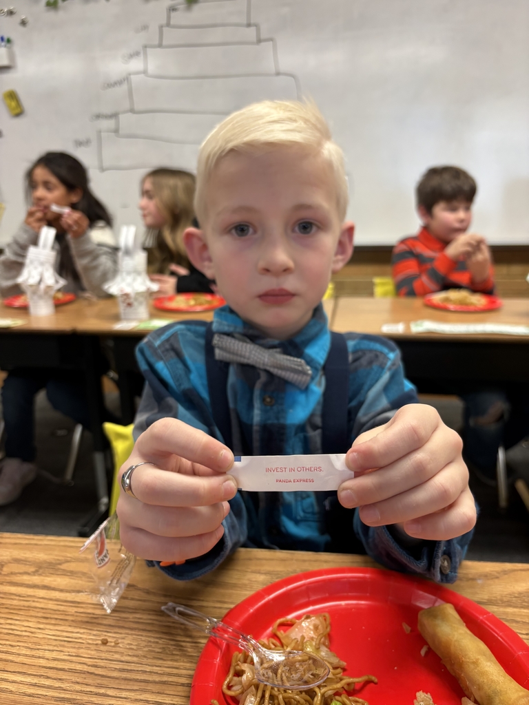 A young boy with blonde hair wearing a blue plaid shirt and a grey bowtie sits at a classroom desk. He holds up a small white fortune cookie slip that reads "INVEST IN OTHERS." In front of him is a red plate with lo mein noodles and an egg roll.