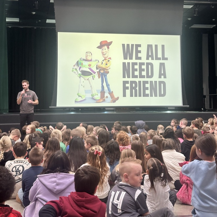 man presenting to large group of students with a sign. we all need a friend. 