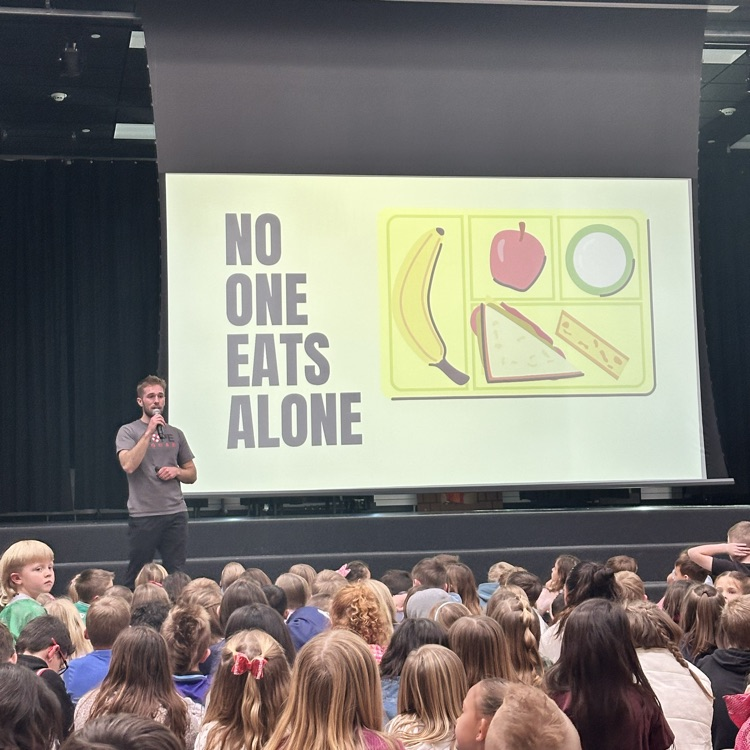 no one eats alone sign with man presenting to large crowd of kids