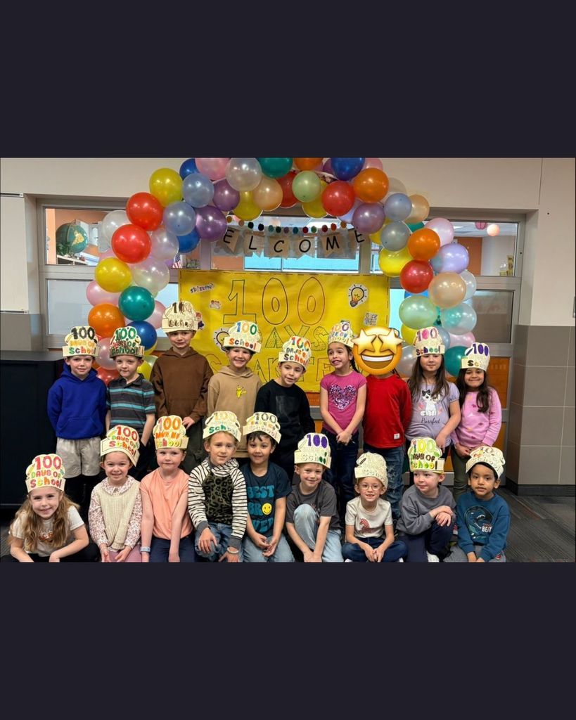 A group of elementary students posing together indoors to celebrate the 100th day of school. Students wear handmade “100 Days of School” hats and stand under a colorful balloon arch in front of a bright yellow display.