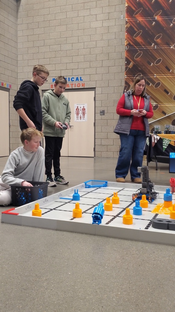 The Grinding Gears Robotics Team competing in a VEX robotics competition at Sunburst Elementary School. Students gather around a competition field, where there robot must pick up and organize an assortment of colorful game pins.