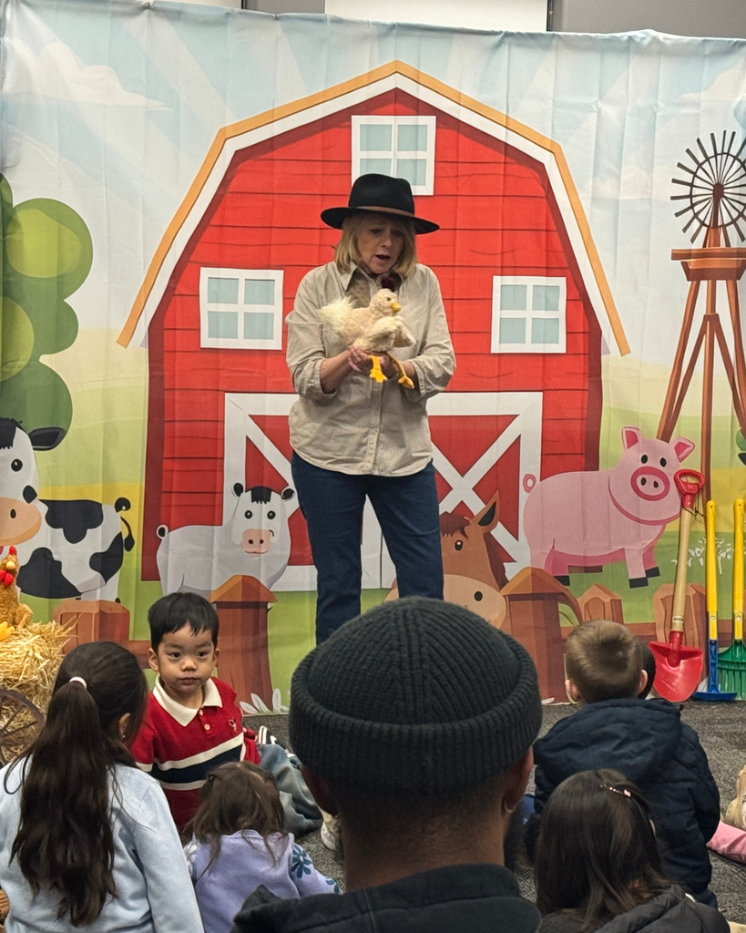 A person holding a duck puppet is talking to students while standing in front of a farm house background.