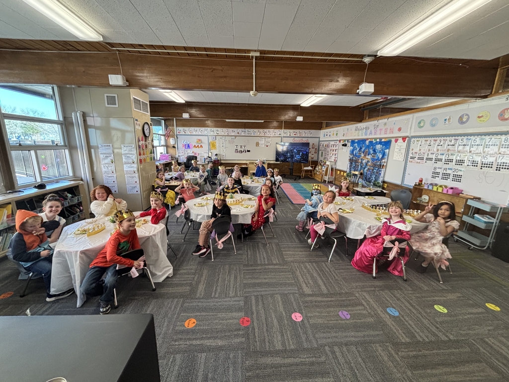 A wide shot of a kindergarten classroom filled with students sitting at round tables with white cloths. Most children are wearing gold paper crowns and "fancy" clothes while smiling for the camera.