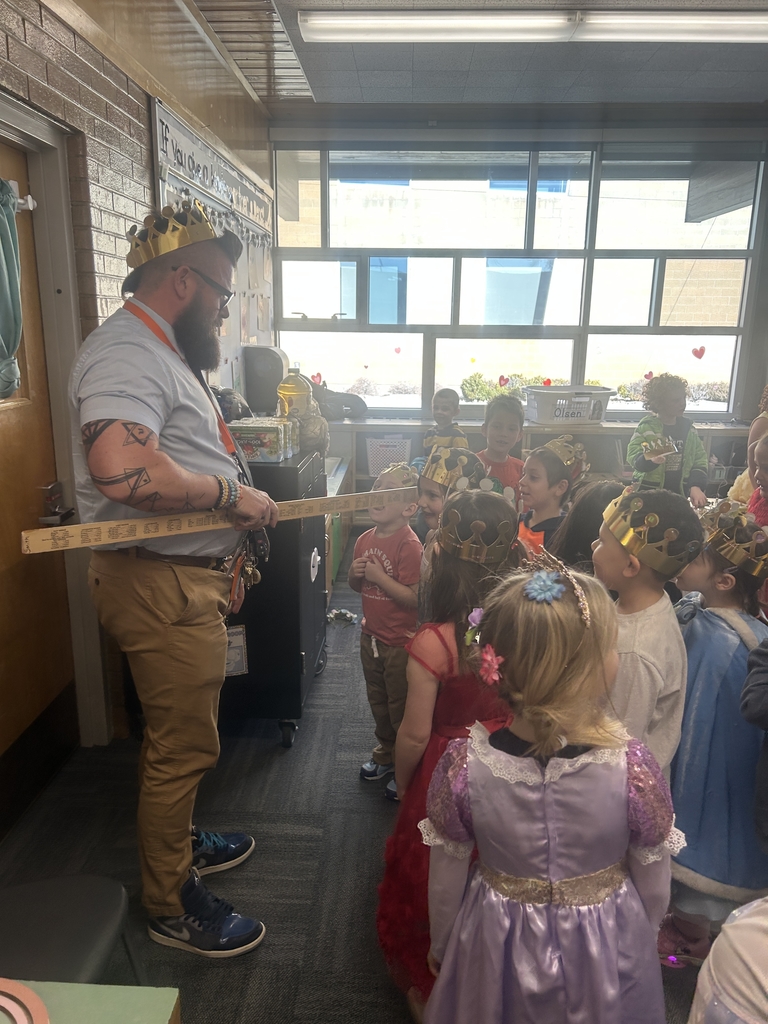 A bearded man (the principal) wearing a crown and a lanyard "knights" a young student by holding a long wooden ruler over their head as other students watch.