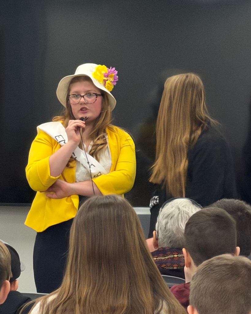 A student in a yellow blazer and white flower hat performs to her peers.