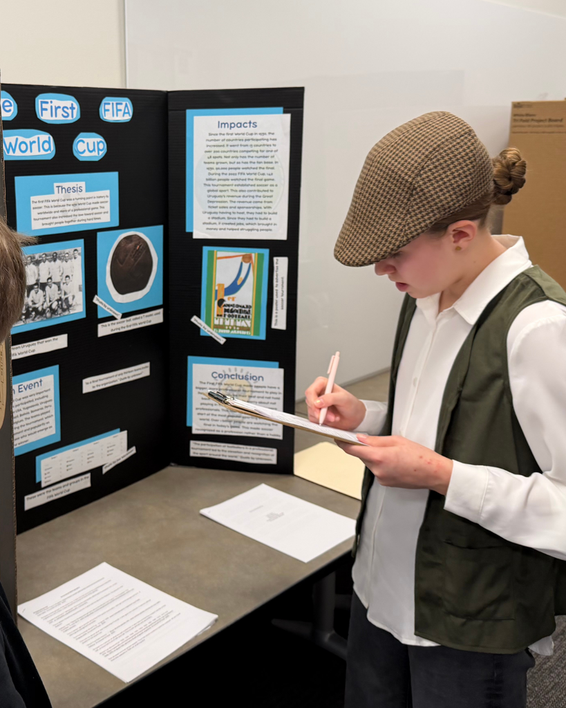 A student in a hat writes on a clipboard. There is a board that says "The First FIFA World Cup"