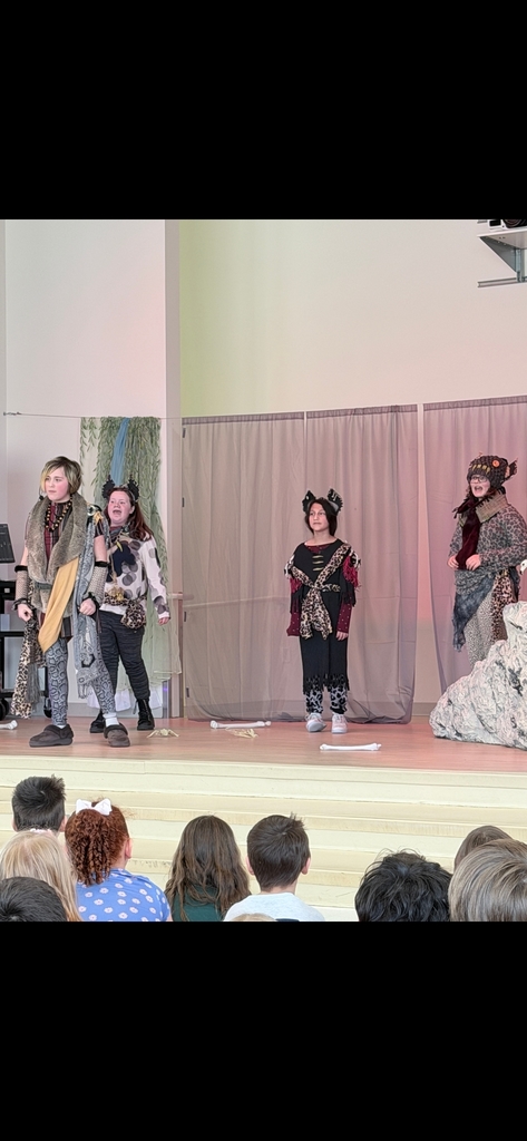 Four students dressed as hyenas perform on stage. They wear grey, black, and leopard print clothing with fuzzy black ear headpieces. The stage features a large grey rock prop and a purple-lit curtain backdrop