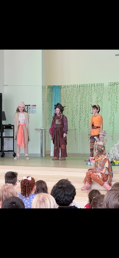 Three students stand on a stage in front of a green leafy backdrop. One is dressed in tan robes and a pink sash (Timone), the middle student wears a dark brown costume with tusks on their head (Pumbaa), and the third wears an orange shirt and animal ears(Simba). A fourth student in a giraffe costume sits to the side
