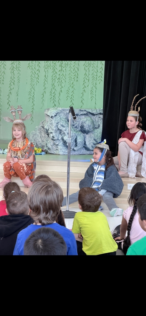 Three elementary students in Lion King costumes sit on a stage during a performance. One student wears a tall, beaded giraffe headpiece; another wears a blue bird headpiece (Zazu); and the third wears a tall, tan headpiece. They are seated behind a microphone, facing an audience of students.