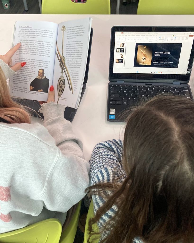Two students compare information from a book and a laptop while working on a presentation about Galileo. One student reads while the other edits slides.