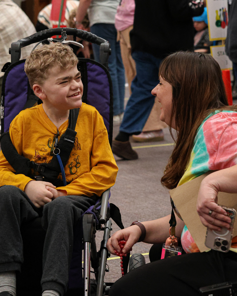 A student in a wheelchair smiles at a teacher who is kneeling at his eye-level.