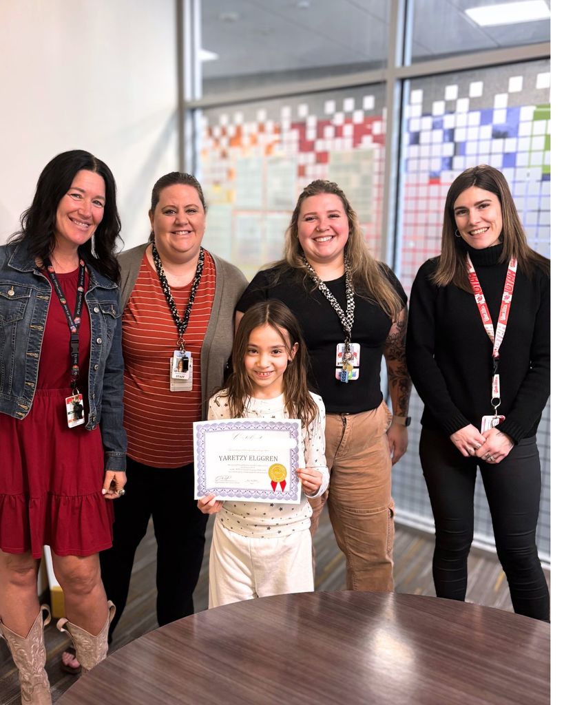A young girl stands in front of four smiling women in a school office setting, holding a certificate with a gold seal and red ribbon. The adults, wearing staff lanyards and badges, stand behind her posing for the photo. The group is in front of large windows with colorful paper displays visible in the background. A round wooden table is in the foreground.
