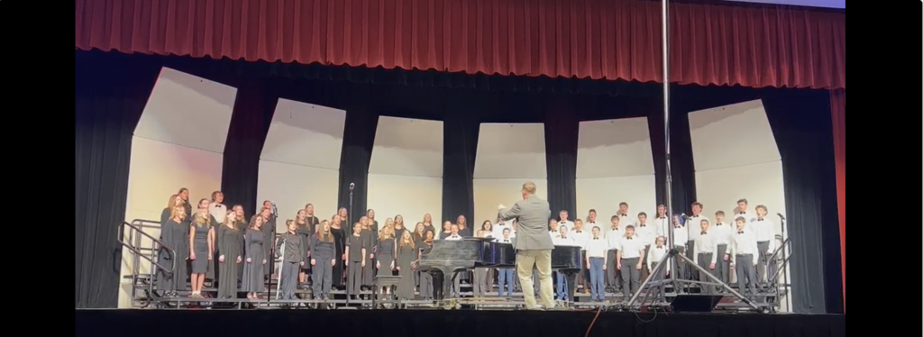 image of students standing on a riser singing, with a director leading the music.