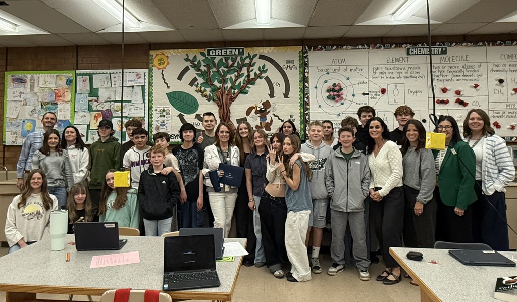 A group of students and teachers stand together in a classroom, smiling for a group photo. Behind them, colorful science-themed posters and a large mural of a tree labeled “GREEN” decorate the wall. Several students hold certificates or papers, and laptops are visible on the tables in the foreground.