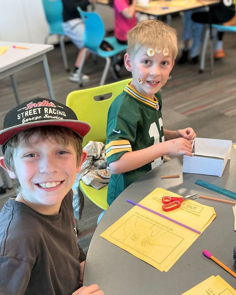 Two third grade students sit at a classroom table working on a STEM activity where they design and build small houses meant to withstand strong winds.