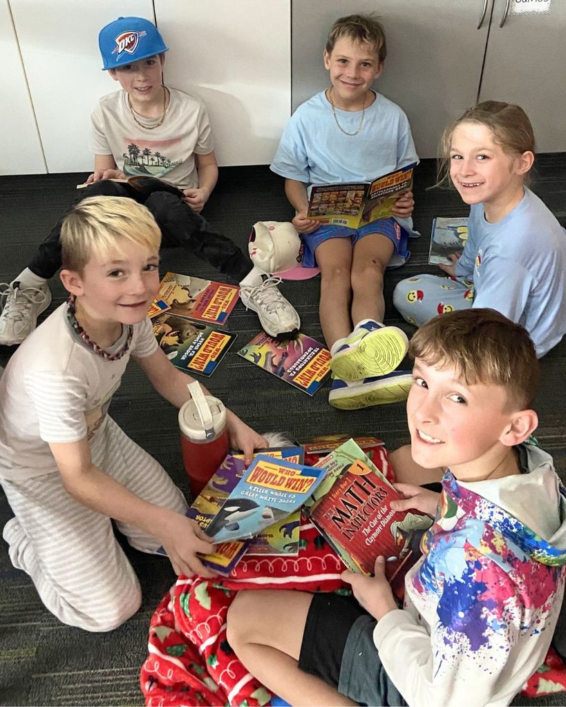 Several elementary students sit in a circle on the floor holding books and smiling at the camera during a Reading Week reading activity.