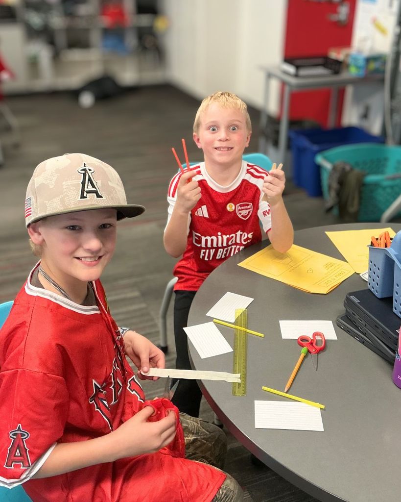 Two students smile at the camera while building small model houses using classroom materials as part of a third grade science lesson about wind and weather.