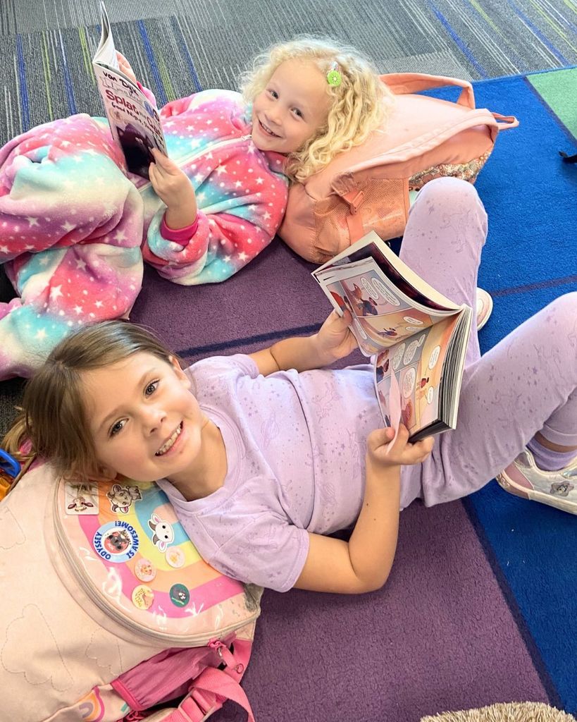 Two elementary students sit on a colorful classroom rug reading books together during Reading Week. One student is wrapped in a rainbow blanket while smiling at the camera, and the other student lies on a backpack while reading a graphic novel.