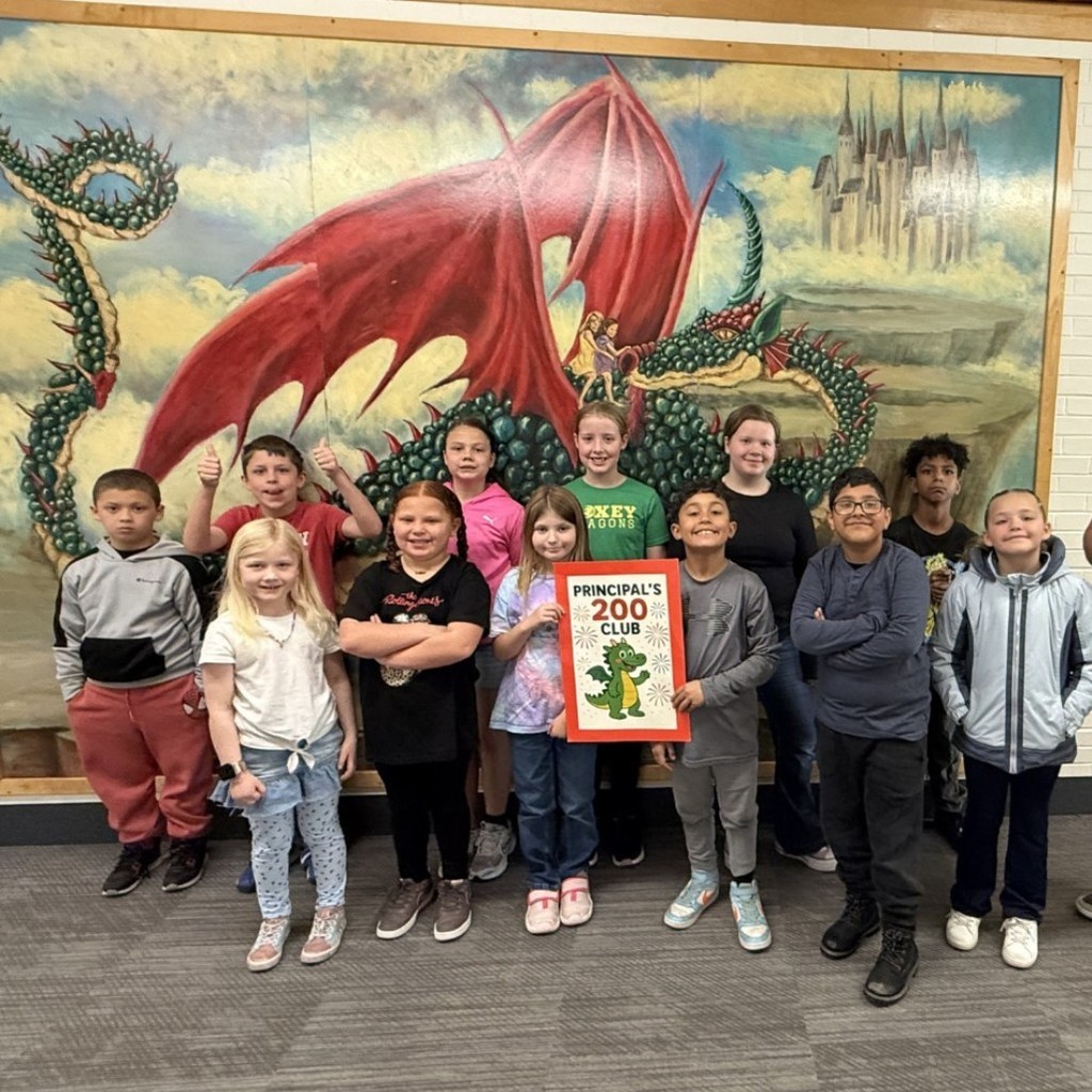 ​A group of eleven elementary students pose proudly in front of a dragon mural. A boy in the center holds an orange sign that reads "Principal’s 200 Club" with a cartoon dragon illustration. A male teacher stands to the right, wearing a yellow shirt and maroon pants, giving a double thumbs up. The students are smiling and posing with arms crossed or thumbs up.