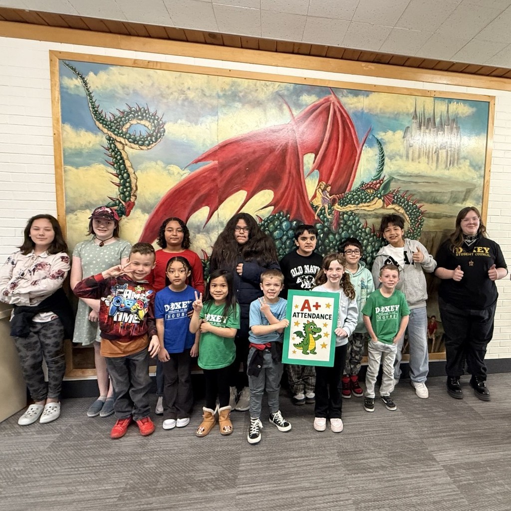 ​A group of thirteen elementary school students of various ages stand in front of a large, colorful mural of a red and green dragon. A girl in the center holds a bright green sign that reads "A+ Attendance" featuring a small cartoon dragon. To the right, a smiling male teacher with a beard and glasses gives two thumbs up. The background includes a "Faculty & Staff" directory board on a white brick wall