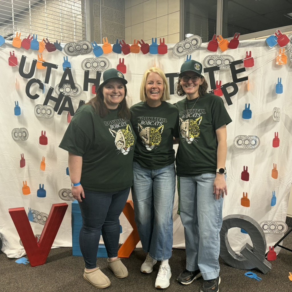 Three women in Bountiful Bobcats shirts pose in front of a VEX IQ photo backdrop.