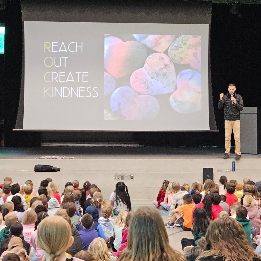 Tom Ballard speaks on stage during a school assembly as students sit on the gym floor watching. A large screen behind him shows colorful heart rocks and the words “Reach Out Create Kindness.”