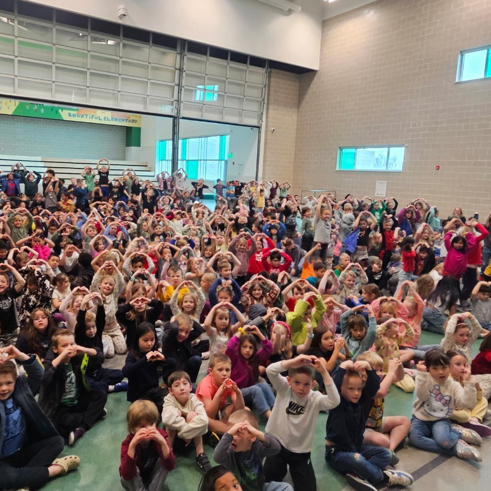 A large group of elementary students and staff gather in the school gym, smiling and making heart shapes with their hands during the Hope Week kickoff assembly.