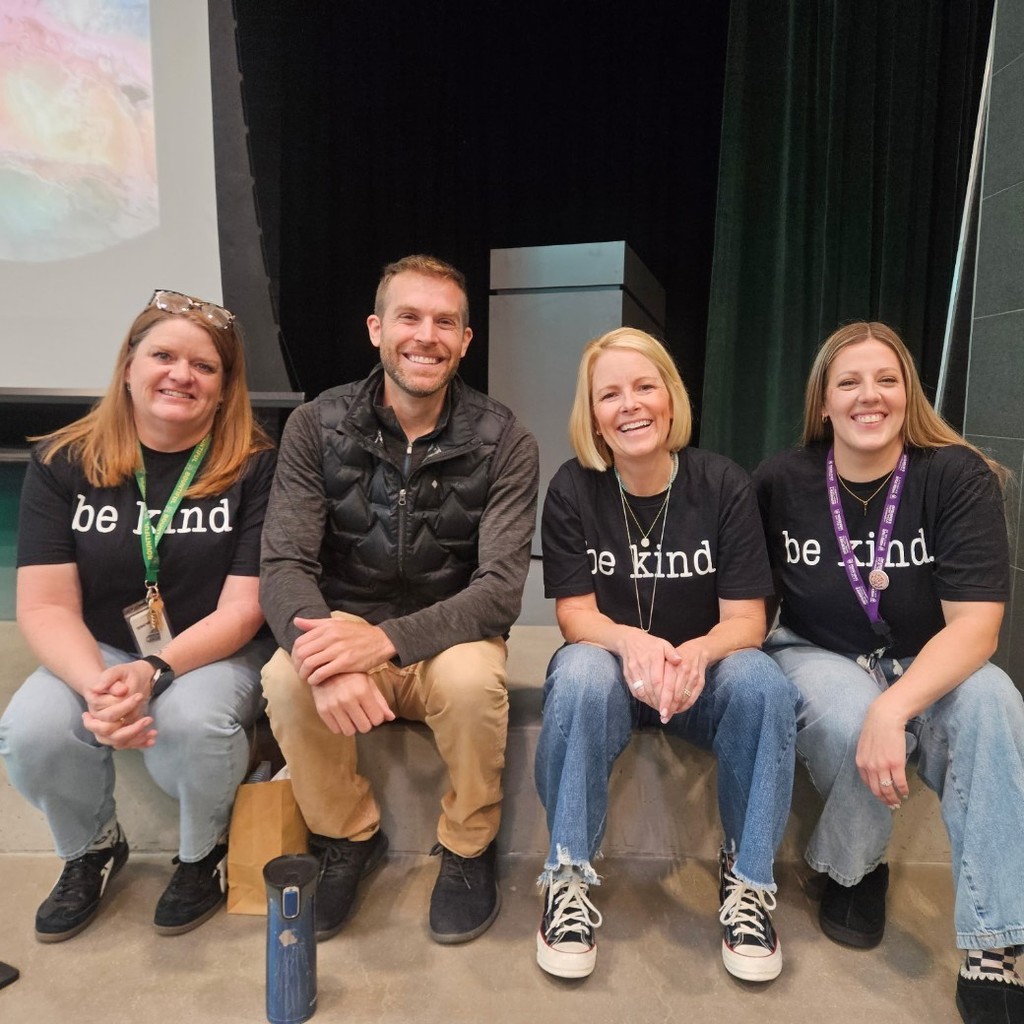 Four adults, including Tom Ballard, sit smiling together on the edge of the school stage. Three staff members wear black “be kind” shirts, and the projection screen behind them shows part of a heart rock image.