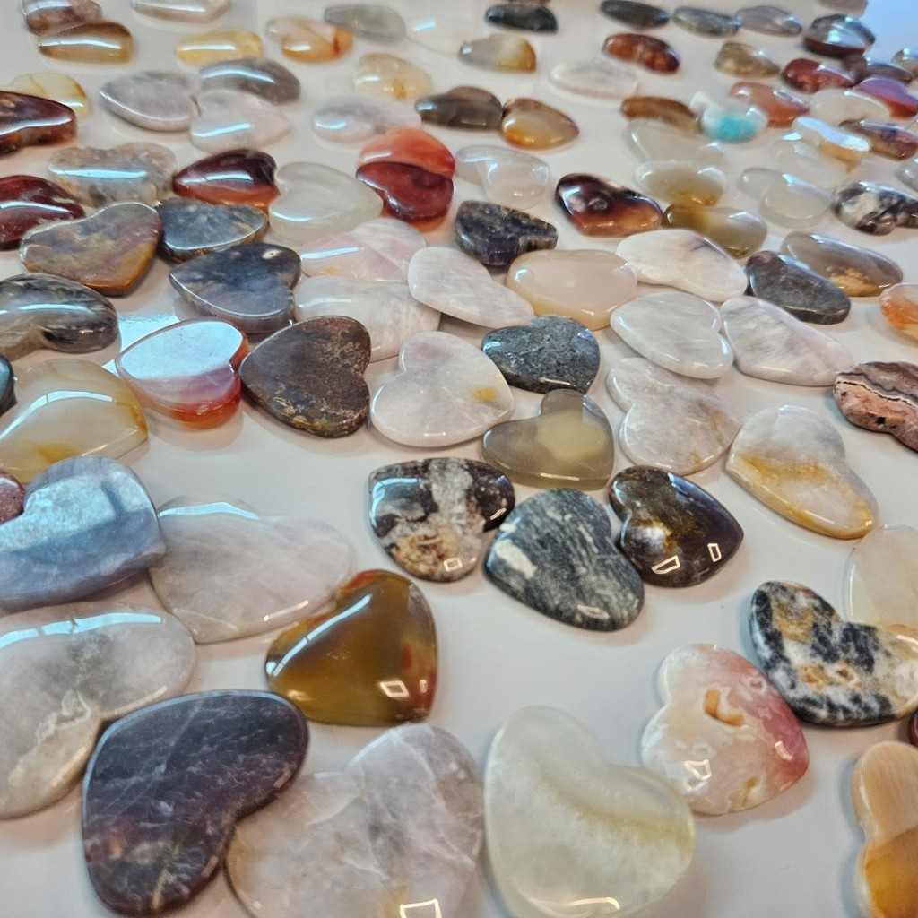 A close-up view of dozens of polished heart-shaped stones arranged on a table in shades of cream, pink, brown, gray, and amber.