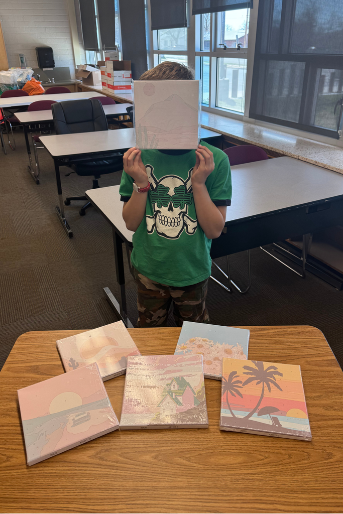 Student in a classroom holding a small canvas painting kit in front of their face while standing behind a table with several painting kits spread out, choosing a prize for their family reading activity.