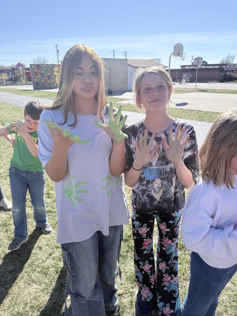 Two girls pose outside with vibrant green powder covering their palms and staining their clothes after a Multicultural Club activity.