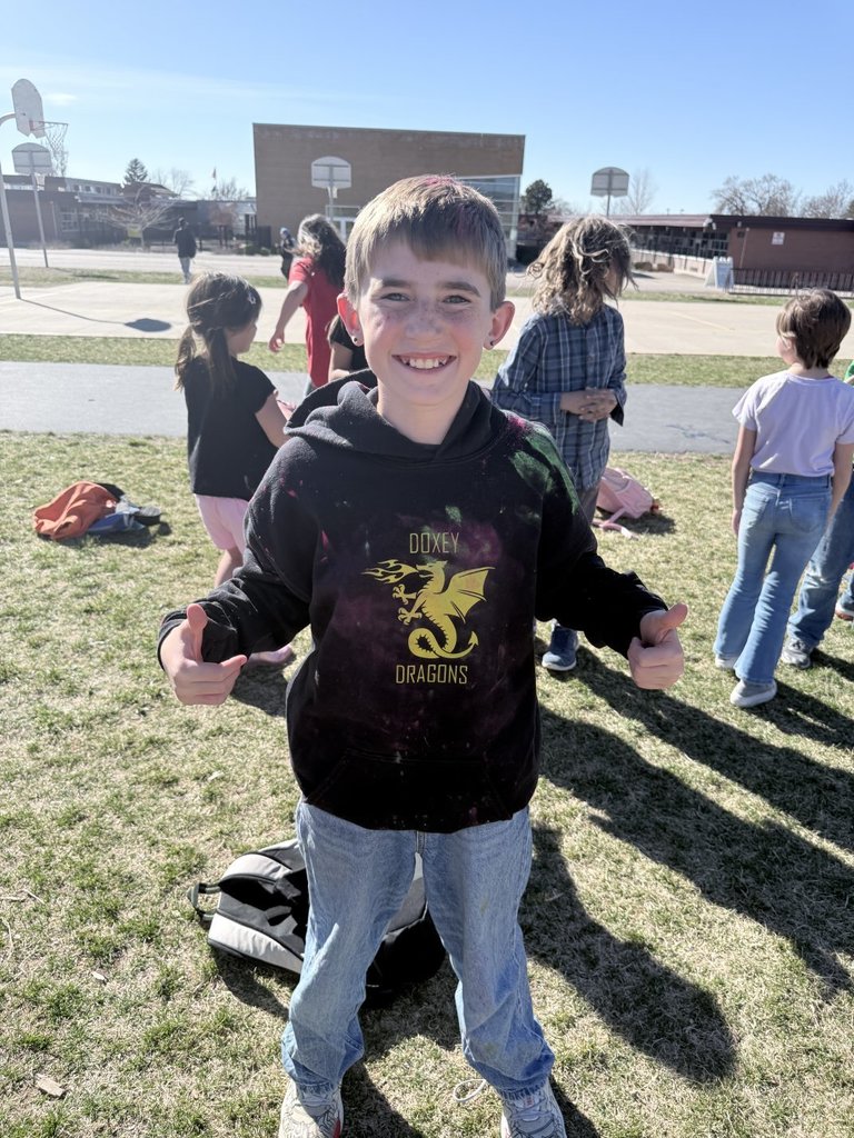A smiling boy wears a black "Doxey Dragons" school hoodie that is speckled with various colors of Holi powder.