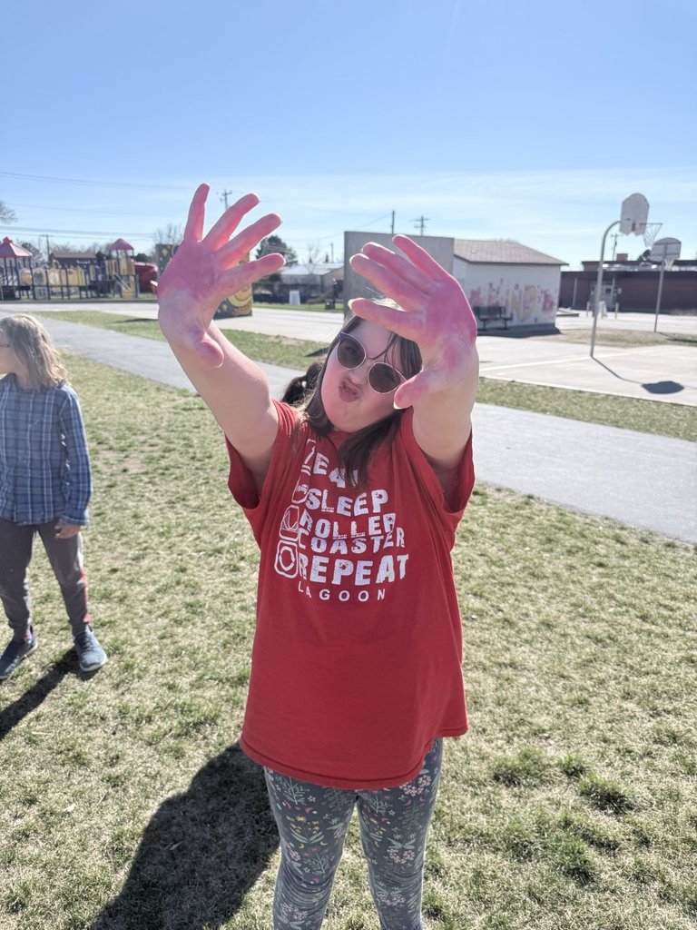 A student in a red t-shirt and sunglasses makes a playful face while holding up her hands, which are stained with bright pink powder.