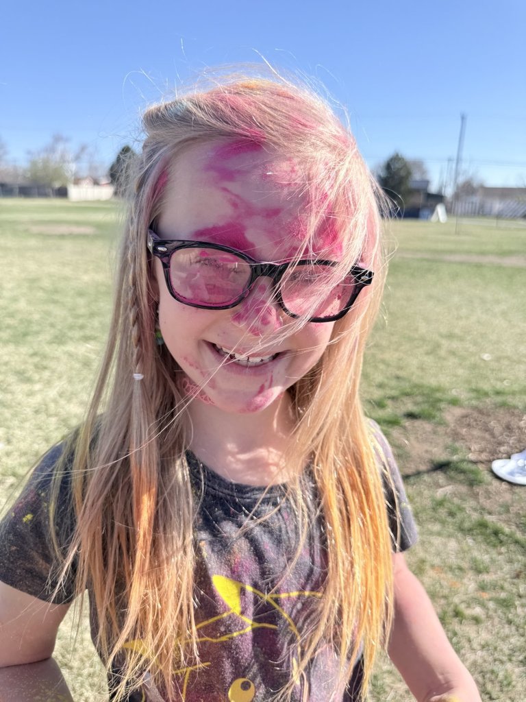 A close-up of a blonde girl with glasses, her face, hair, and frames completely covered in vibrant pink Holi powder as she smiles.
