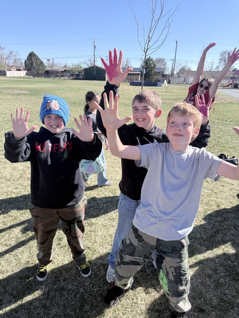 Three boys wave enthusiastically at the camera on the school field, with one boy showing his palms covered in pink powder.