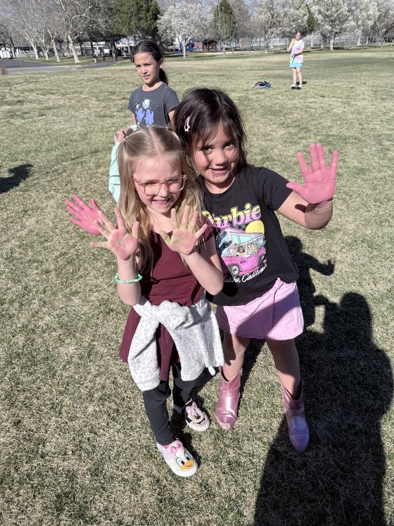 Two young students at Doxey Elementary smile on a grassy field, showing off their hands covered in bright pink and green Holi powder.