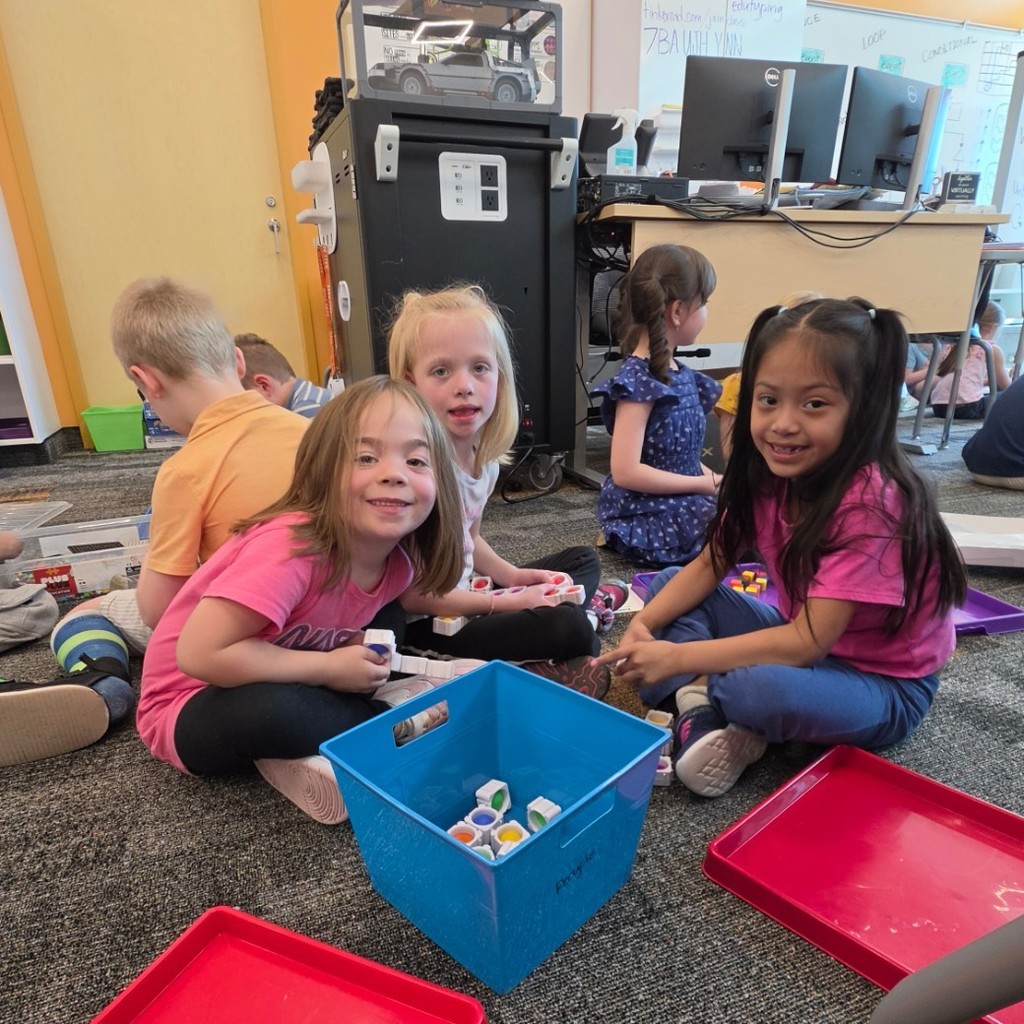 Students sit across the computer lab floor using trays and building materials during a hands-on STEM activity.