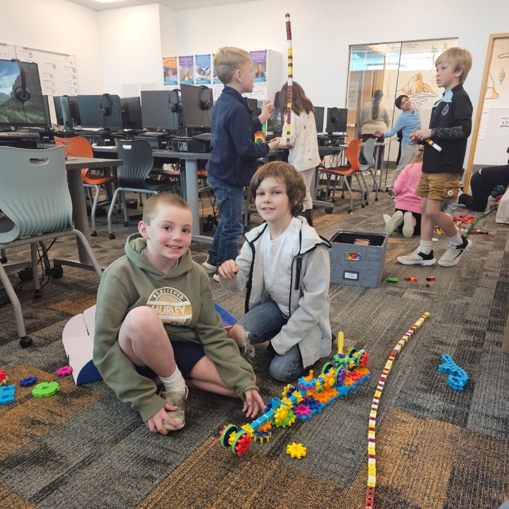 Two students sit on the floor building small vehicles and structures with colorful interlocking blocks.