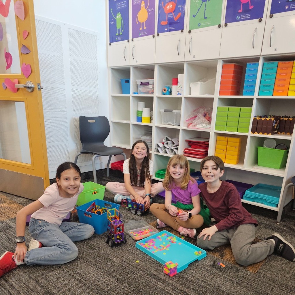 Four students sit together on the floor beside magnetic tile builds and a tray of small colorful cubes.