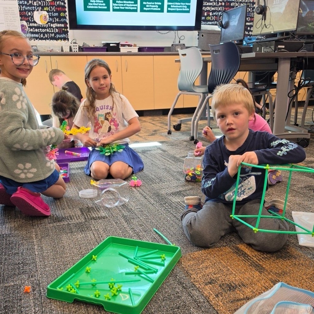 Three students build with green rods and connectors on the computer lab floor, and one student holds a cube-shaped frame.