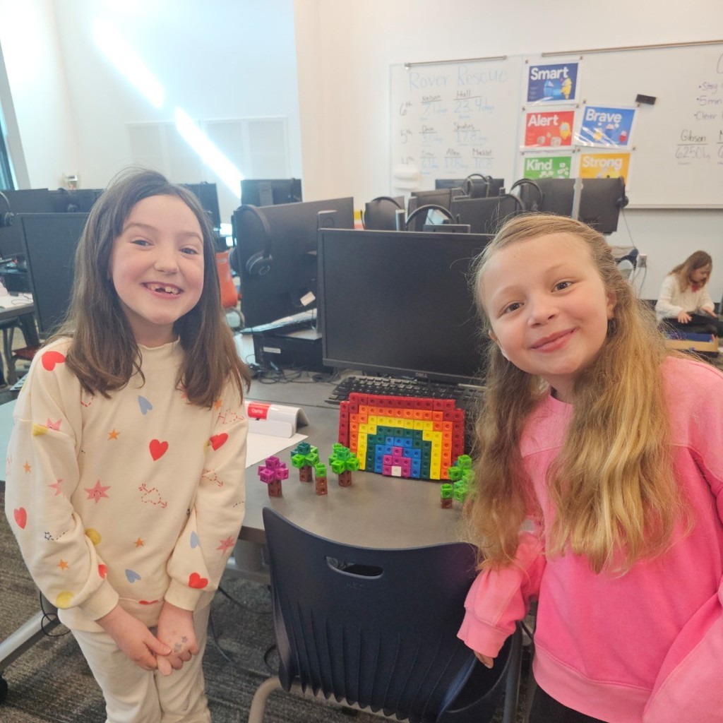 Two students stand beside a rainbow built from colorful interlocking cubes on a table in the computer lab.