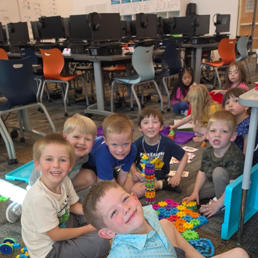 Six students sit on the computer lab floor building with large colorful gears and connectors while other students work in the background.
