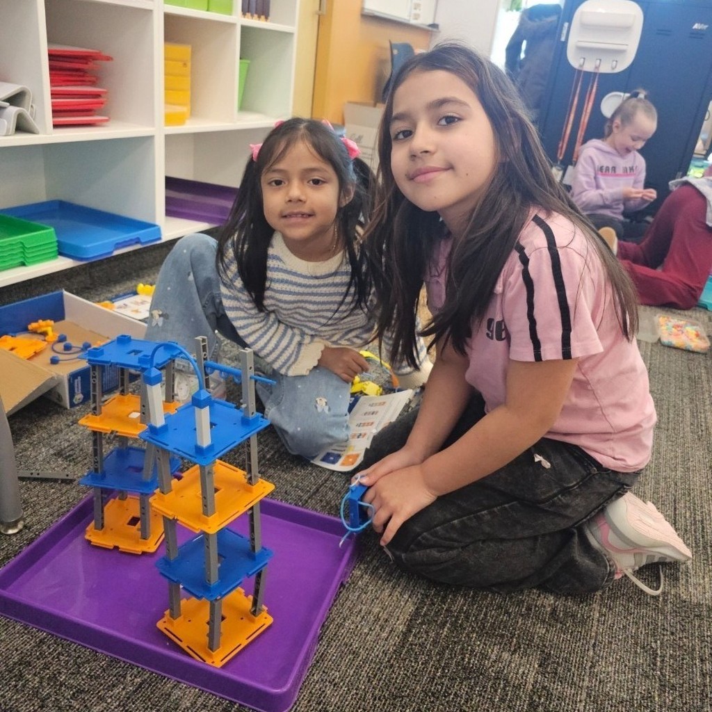 Two students kneel beside a tall blue and orange tower built on a purple tray in the computer lab.